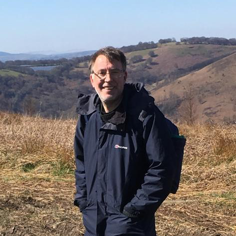 Owen is smiling, wearing glasses. He is on a trek in the outdoors with beautiful hilly landscape in the background.