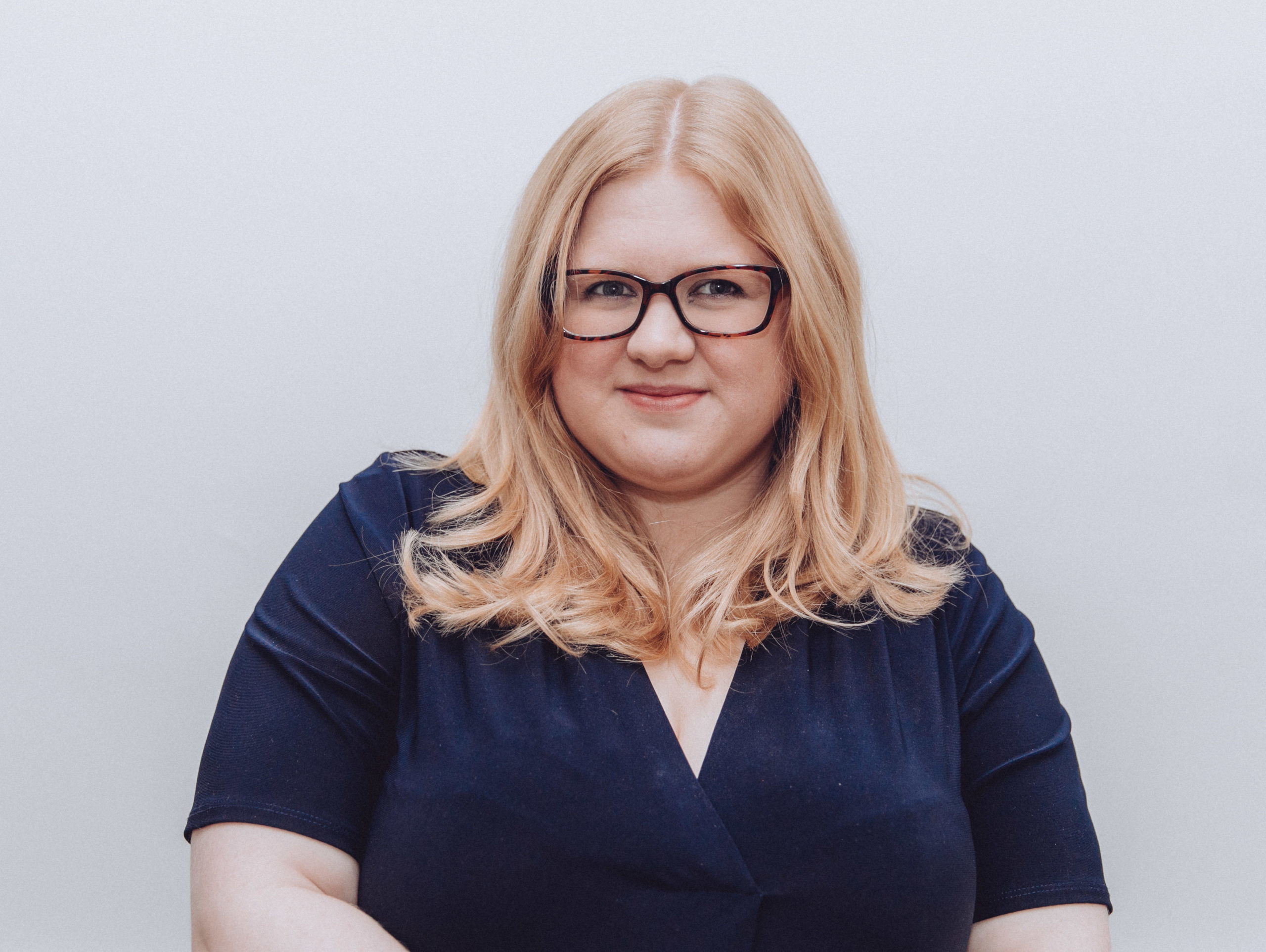 A headshot. Amy is a blonde white plus size woman wearing glasses. She is half smiling at the camera and wears a navy dress