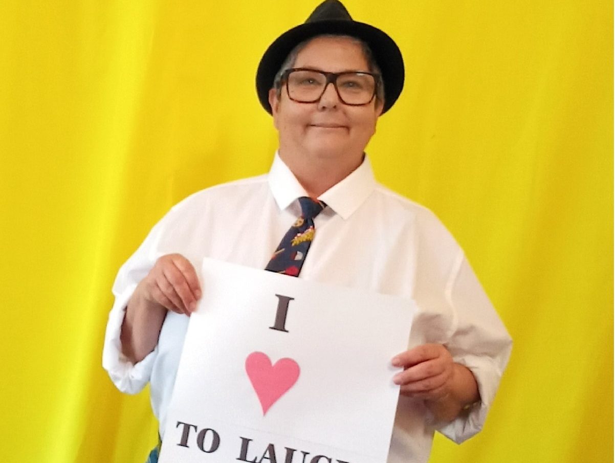 Image is a photo of Angela de Castro smiling at the camera holding a sign which says I love to laugh