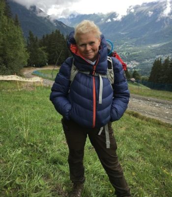Image is a photo of Fi standing in front of a hillside view. Fi is dressed for a long walk with backpack and warm clothes