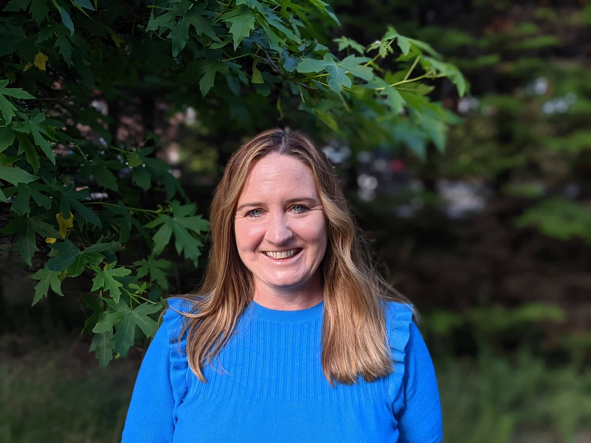 Kirstie Thomas smiling, wearing a blue top with trees in the background.