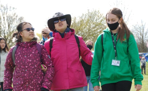 Image is three people walking,. Person in the centre is suported by a sighted guide