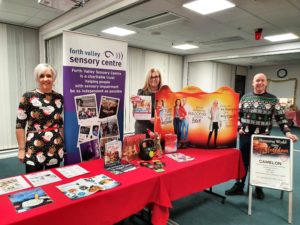 Image is a photo of 3 people standing alongside a display table.  The table features information and posters about Slimming World and Forth Valley Sensory Centre