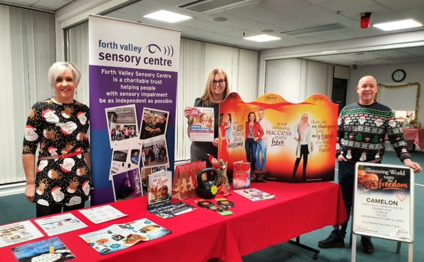 Image is a photo of 3 people standing alongside a display table. The table features information and posters about Slimming World and Forth Valley Sensory Centre
