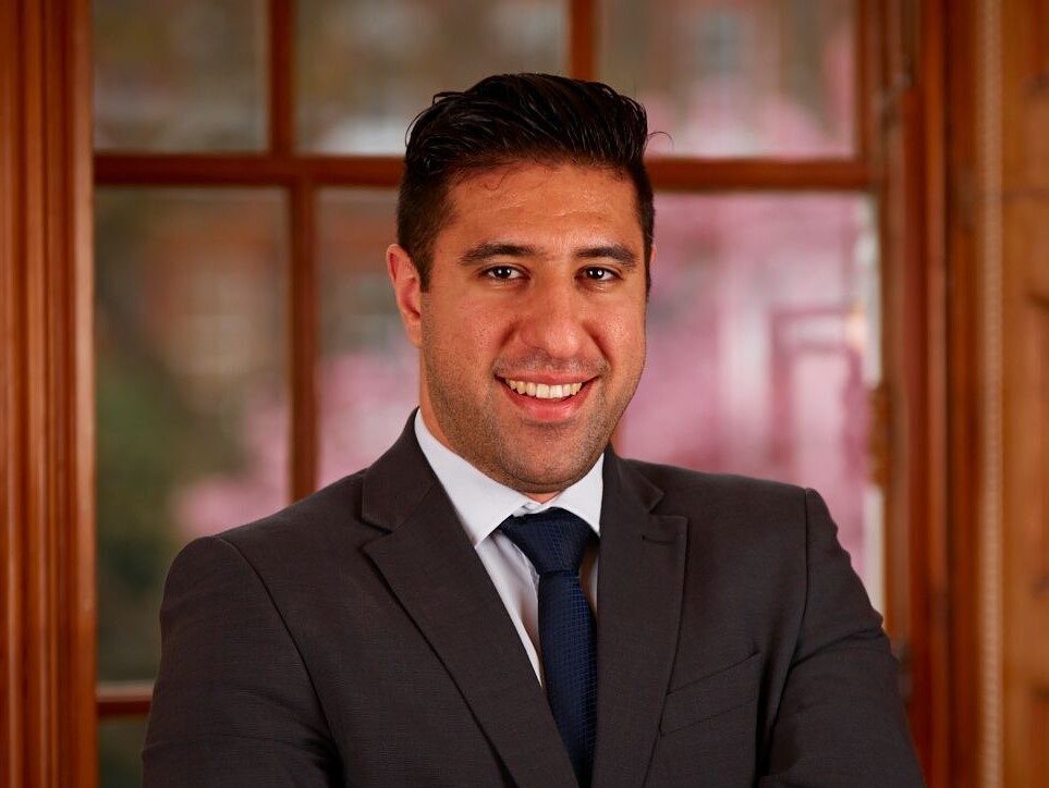 Image is a profile picture of Arash, smiling with his arms folded.  Arash is wearing a dark jacket and tie with a wooden framed backdrop. 