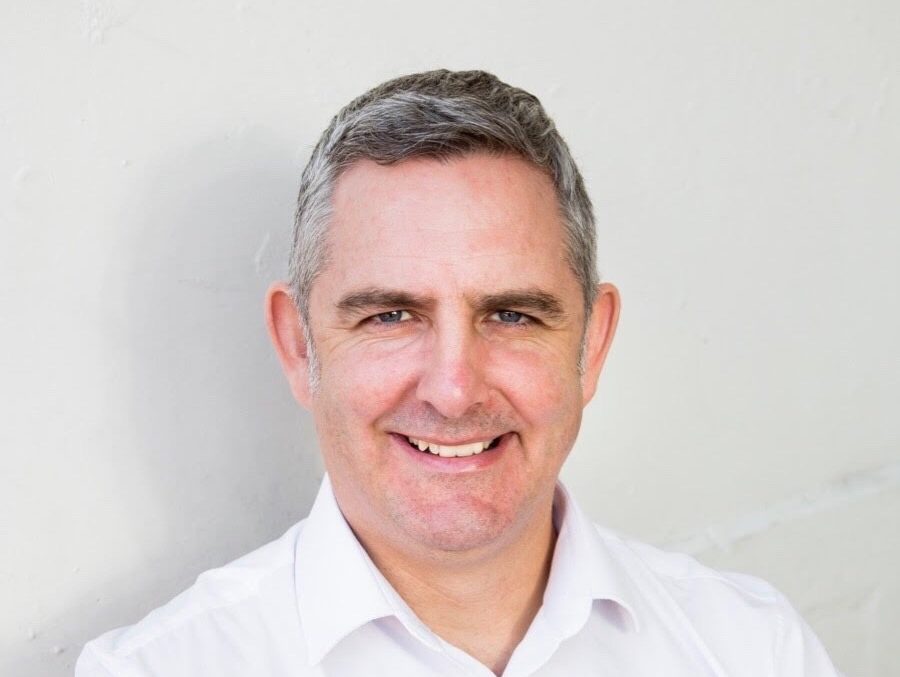 Image is a head and shoulders photo of Tom, against a white background.  Tom is smiling at the camera wearing a white shirt. 