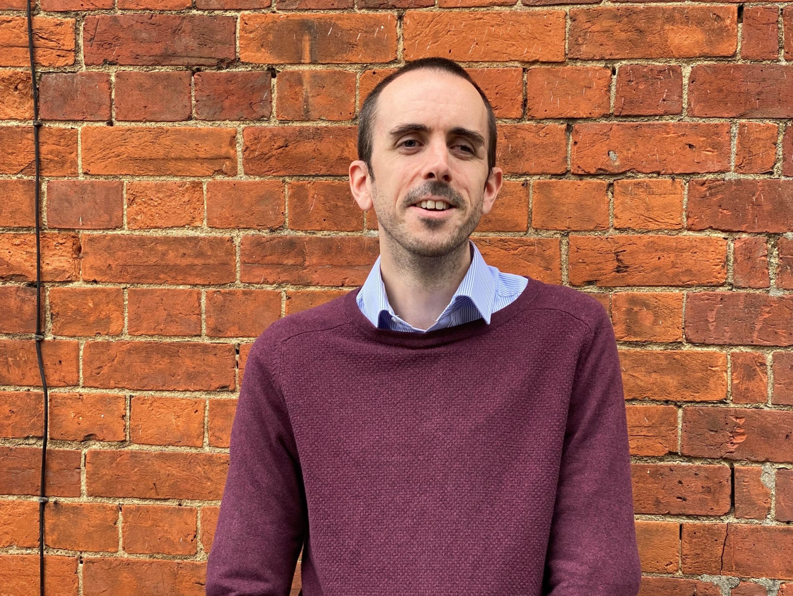 Image is a photo of Mark, smiling at the camera with his arms cross in front.  the image is taken outside with a brick wall background, Mark is wearing a jumper and shirt. 