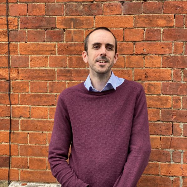 Image is a photo of Mark, smiling at the camera with his arms cross in front.  the image is taken outside with a brick wall background, Mark is wearing a jumper and shirt. 