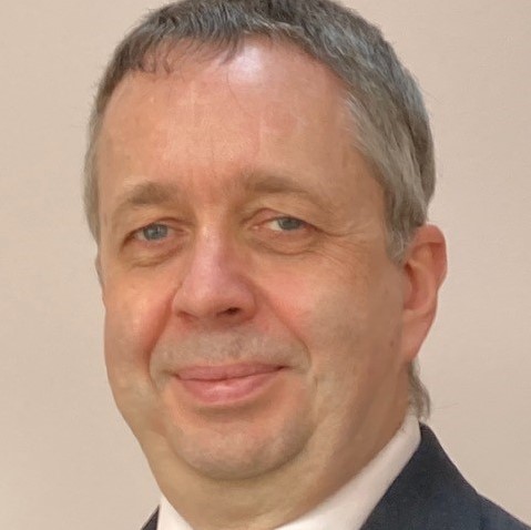 Image is a photo of Philip smiling at the camera, Philips is wearing a jacket, shirt and tie against a cream background.