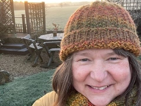 Image a is photo of Susan, taken outdoors with the sun setting behind.  Susan is smiling at the camera wrapped up warm in a hat and scarf. 