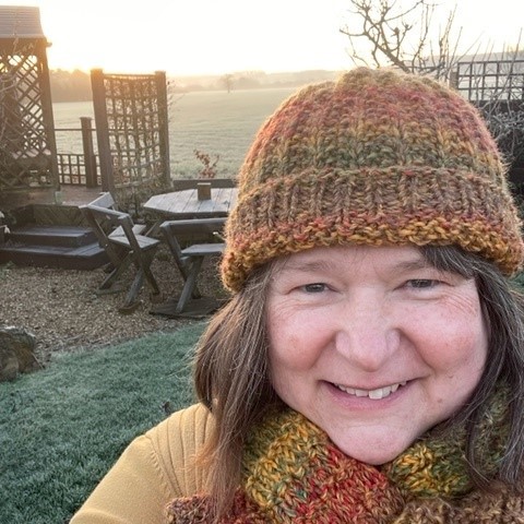 Image a is photo of Susan, taken outdoors with the sun setting behind.  Susan is smiling at the camera wrapped up warm in a hat and scarf. 