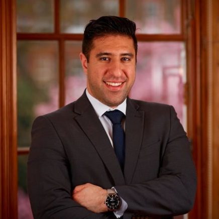 Image is a profile picture of Arash, smiling with his arms folded. Arash is wearing a dark jacket and tie with a wooden framed backdrop.