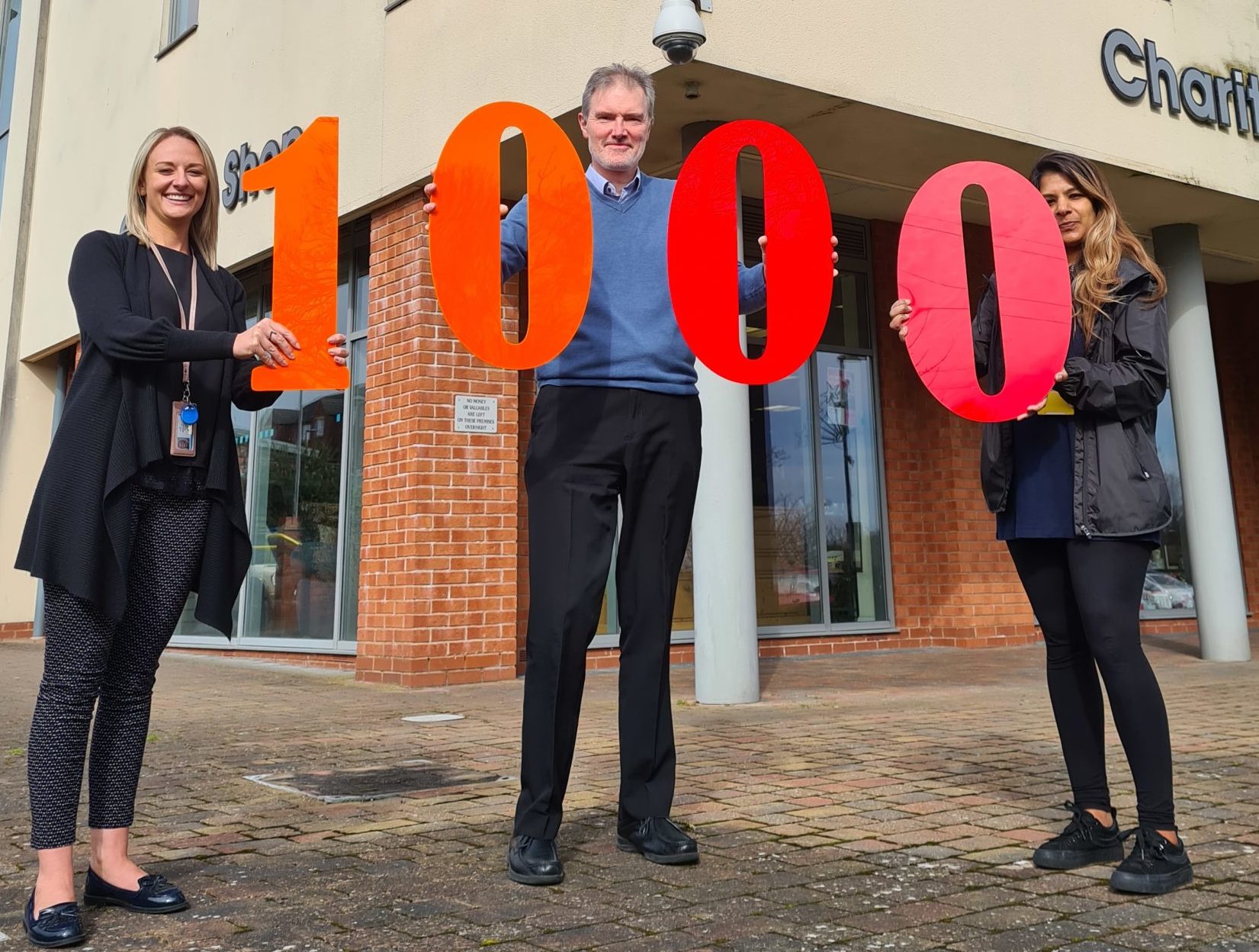 Photo of three of Beacon’s Engagement team, Becky on the left with blond hair, Pete the man in the middle and Meena on the right with long dark hair. They are stood outside the Beacon Centre and are holding up large Perspex numbers which spell out 1000.