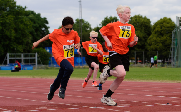 Photo of 4 young children in sportswear running outdoors on an athletic track.