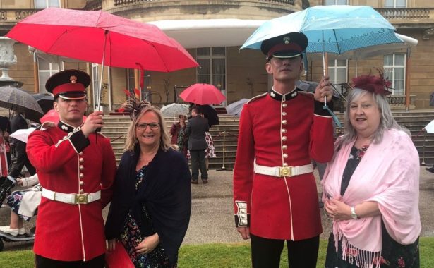 Left to right - Sandra Ackroyd and Angela Gregory at Queens garden party wearing long, flowy dresses, shawls and fascinators, standing under umbrellas held by royal guards.