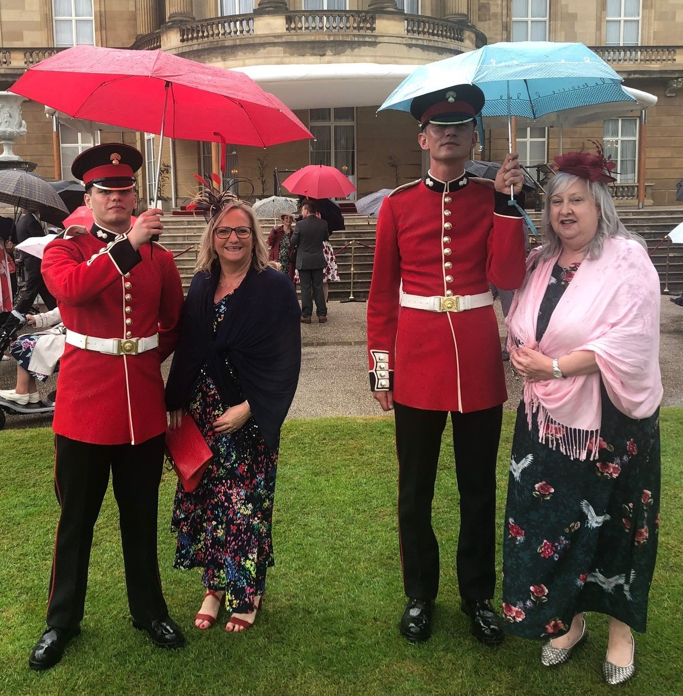 Sandra Ackroyd (left) and Angela Gregory (right) at Queens garden party wearing long, flowy dresses, shawls and fascinators, standing under umbrellas held by royal guards. 