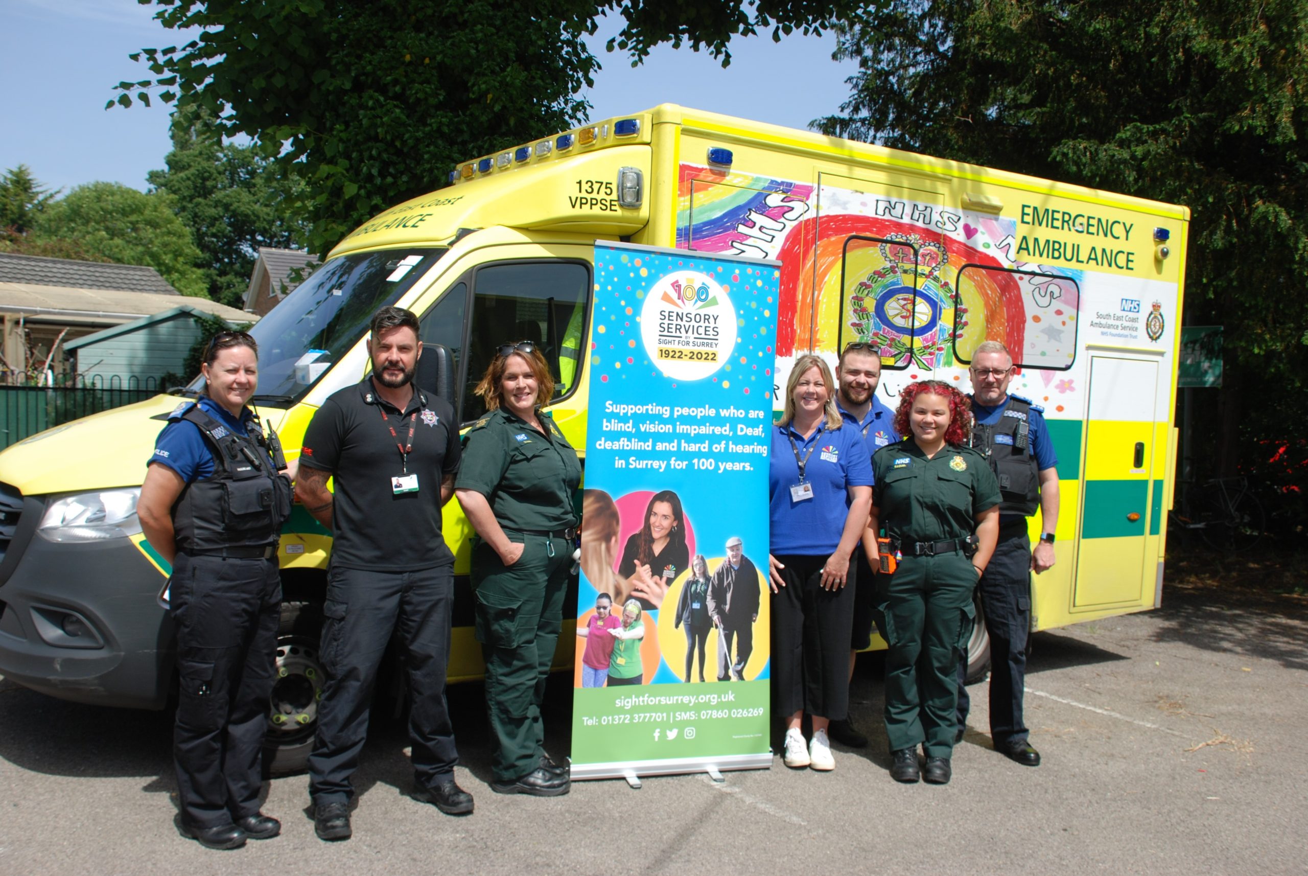 Team 999 BSL (Surrey Police, Surrey Fire & Rescue & South East Coast Ambulance Service and Sight for Surrey staff) standing in front of an ambulance on either side of a Sight for Surrey banner.