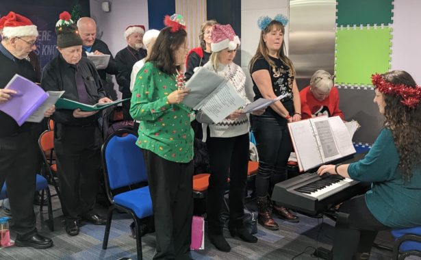 Choir in full swing wearing festive hats and head gear, with a pianist at the front facing the choir
