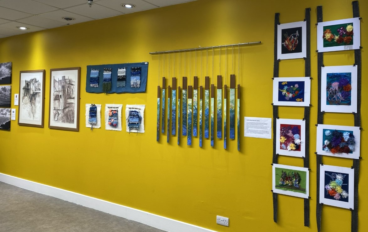 Artwork including some tactile art from fabric and wool displayed against the backdrop of a bright yellow wall.