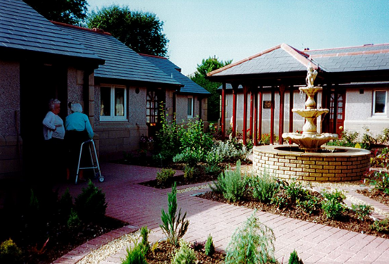 Two female residents (one with a zimmer frame) of Ostley House residential home talking in the garden. There is a fountain in the middle of the garden. 