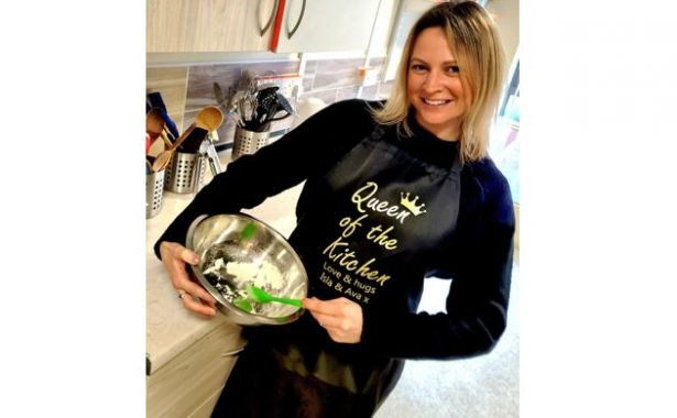 A woman in a kitchen wearing a black apron with a gold crown on it and gold writing "Queen of the kitchen. Love and hugs Isla and Ava x".