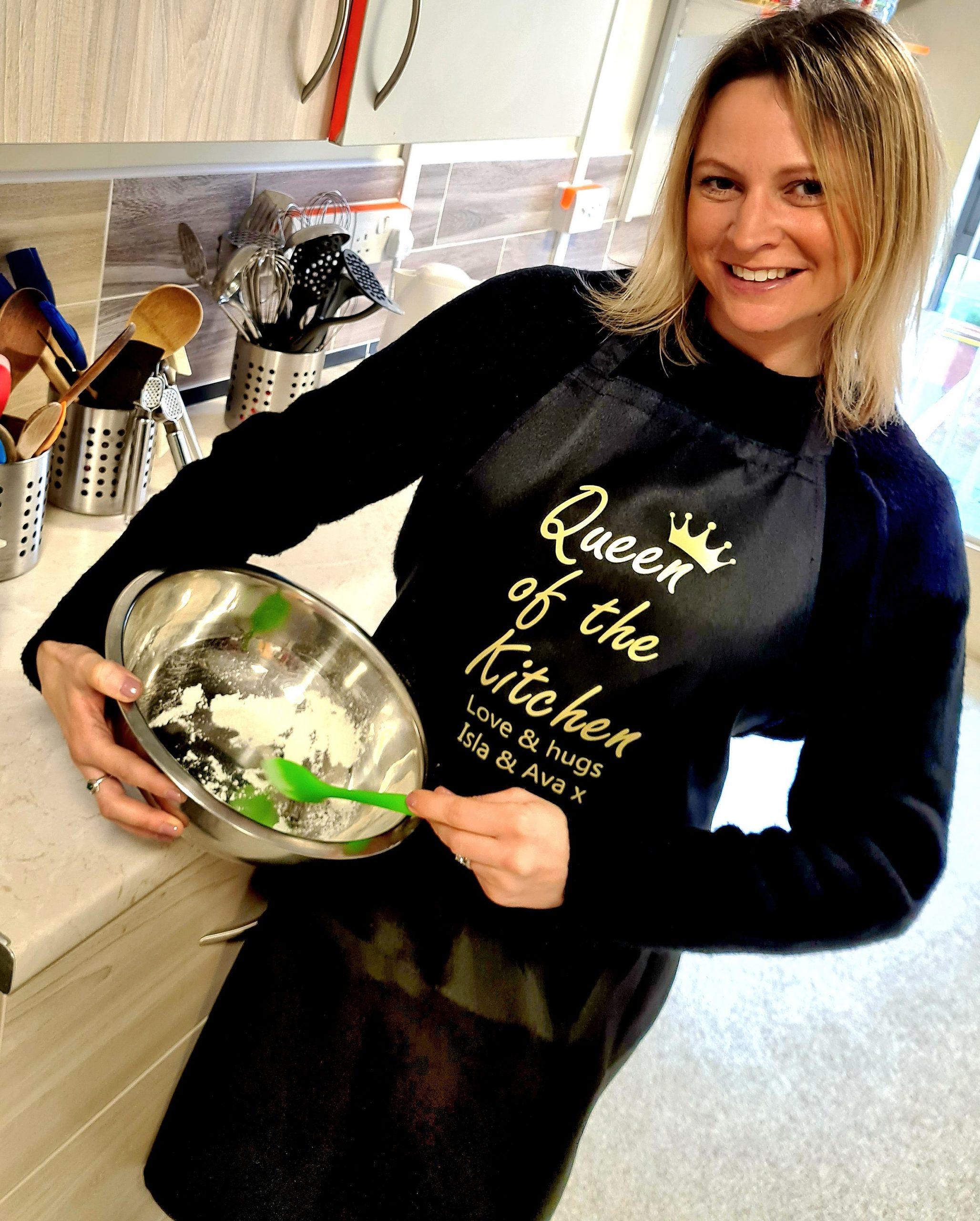 A woman in a kitchen wearing a black apron with a gold crown on it and gold writing "Queen of the kitchen. Love and hugs Isla and Ava x".