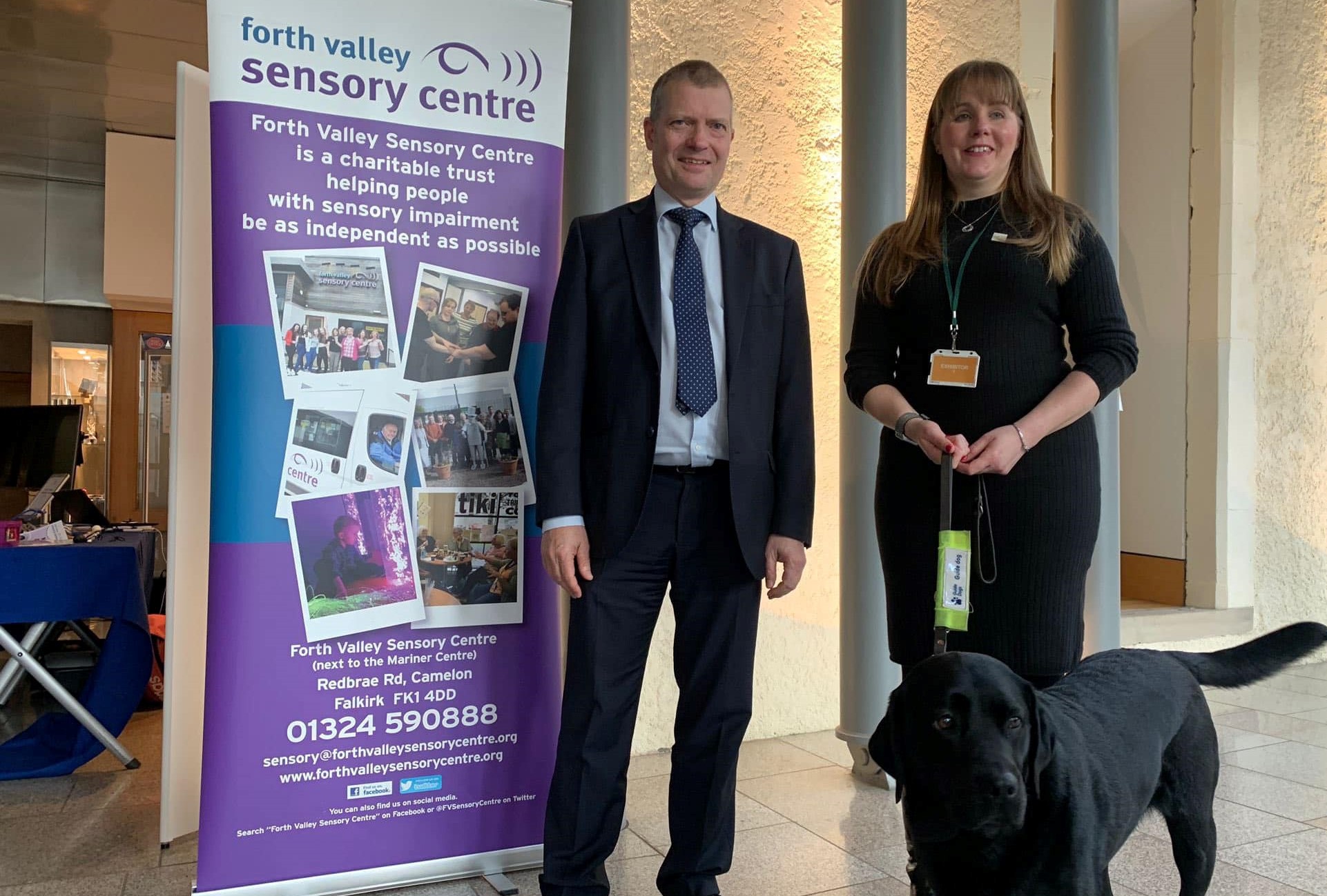 Laura Cluxton with guide dog Sadie and Graham Simpson MSP standing by Forth Valley Sensory Centre exhibition stand.