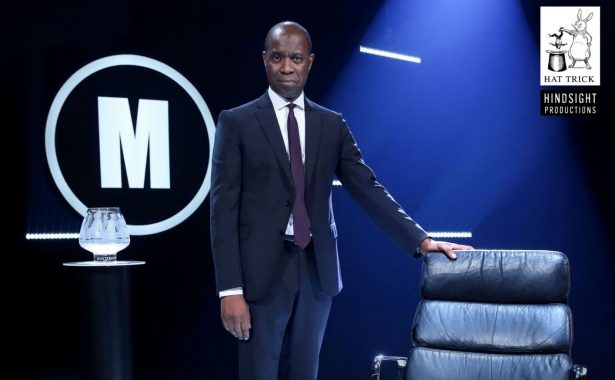 Mastermind Presenter Clive Myrie stands with his hand on the iconic Mastermind Black Chair on his right hand side, and the Mastermind Winner Trophy on his left. Terms and conditions are displayed underneath in white writing on a black background.