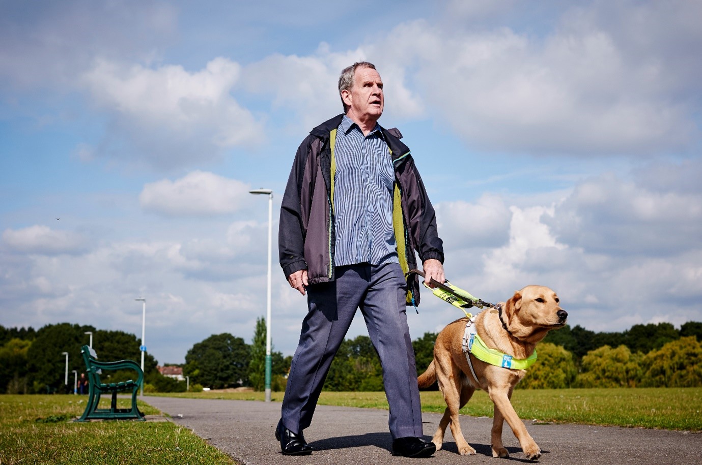 A man walking with a guide dog with grass on either side of the path, trees in the background and clouds in the blue sky.