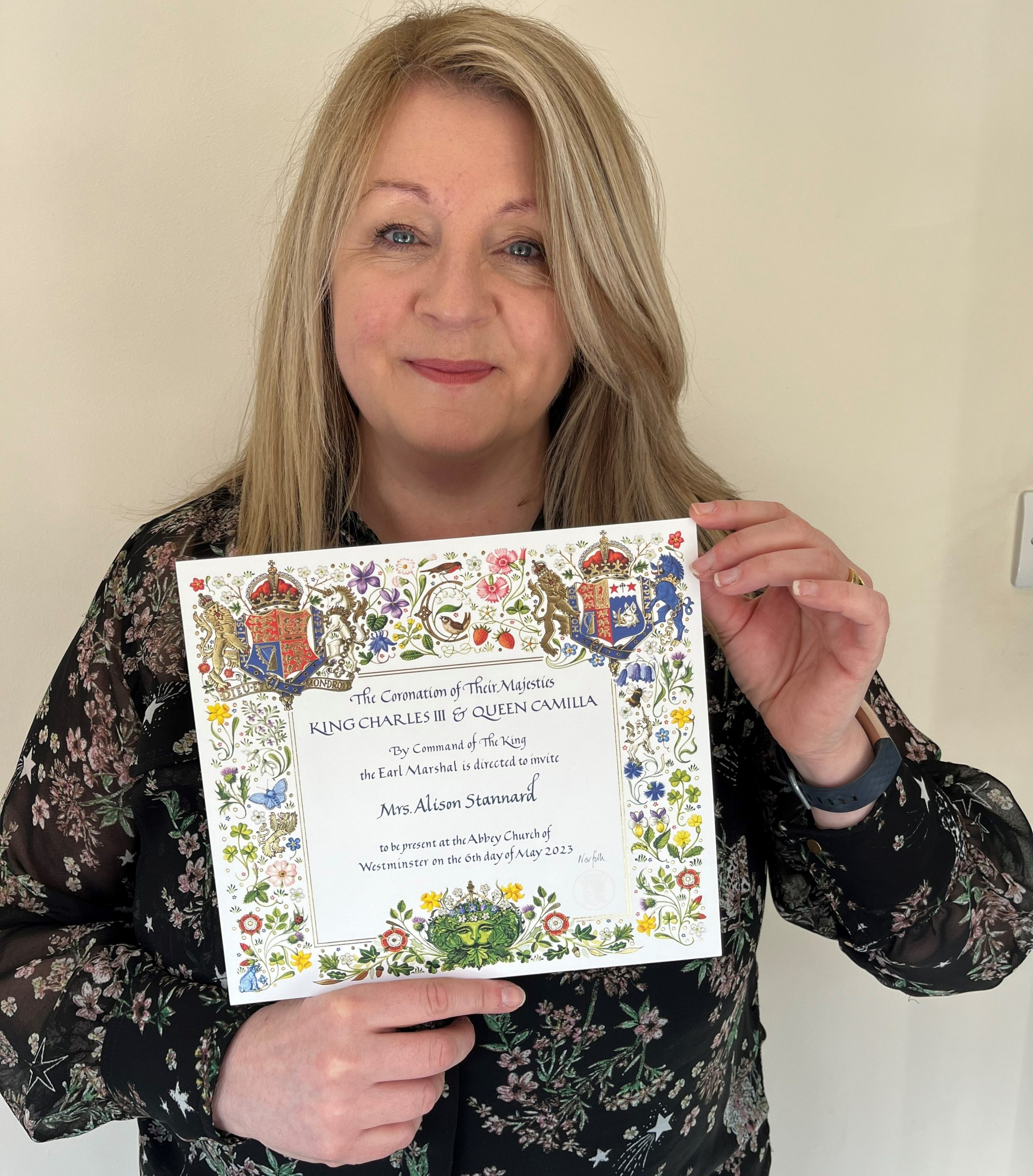 Alison holding up King's Coronation invitation with elaborate border filled with nature - birds, insects, animals, plants and fruit and royal crest in top right and left hand corner.