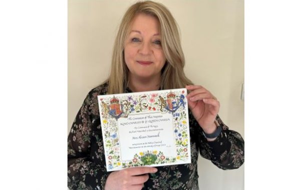 Alison holding up King's Coronation invitation with elaborate border filled with nature - birds, insects, animals, plants and fruit and royal crest in top right and left hand corner.