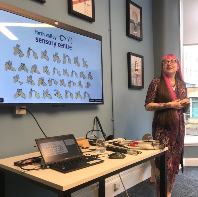 Hannah Wilson giving a sensory awareness training session. On the screen is sign language with the heading Forth Valley Sensory Centre logo at the top. There are canes on the table as well as one leaning against the wall.