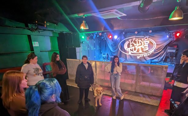 King Tut's venue with logo on the stage. A guide dog owner with a guide dog is in the centre of the floor and staff are standing around them.