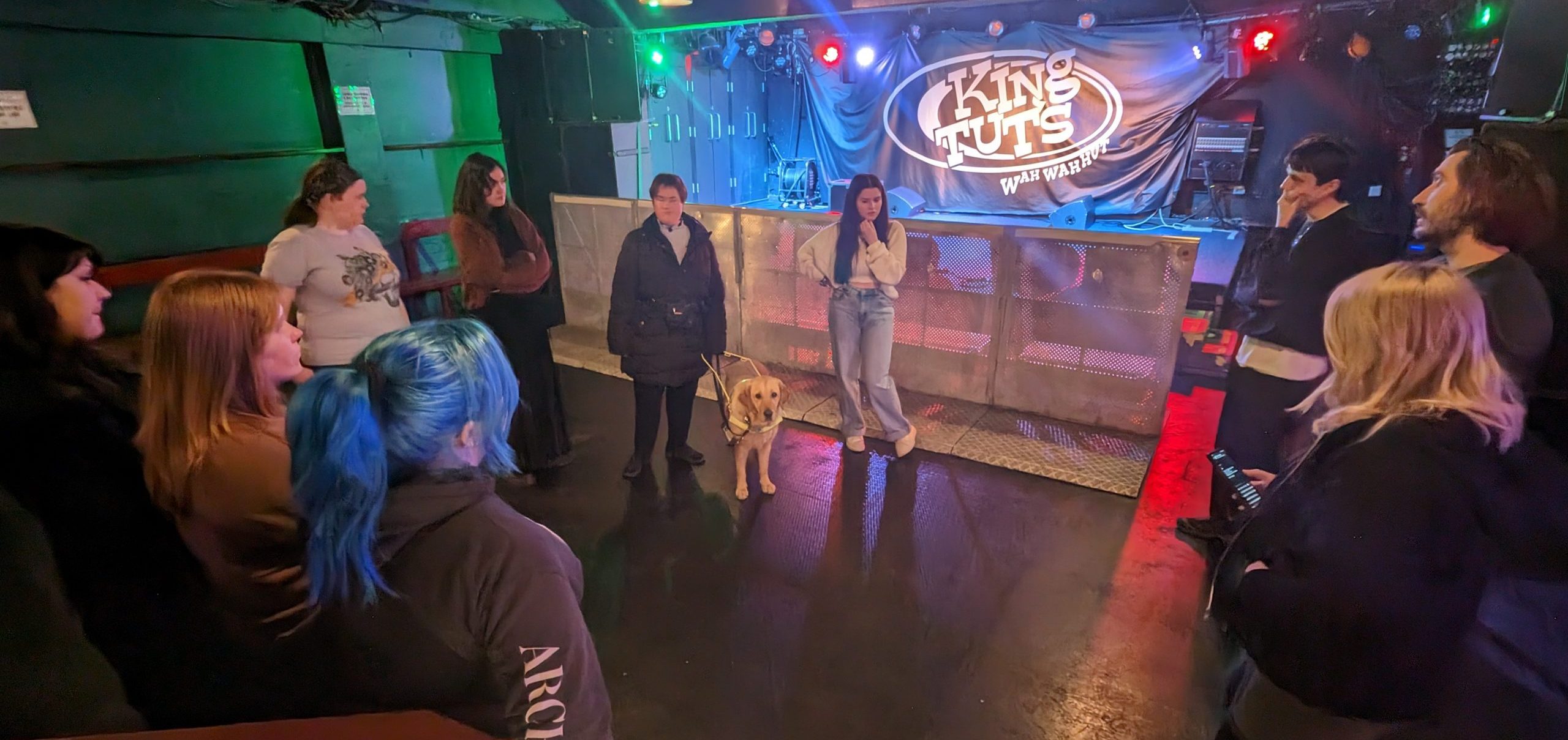 King Tut's venue with logo on the stage. A guide dog owner with a guide dog is in the centre of the floor and staff are standing around them.