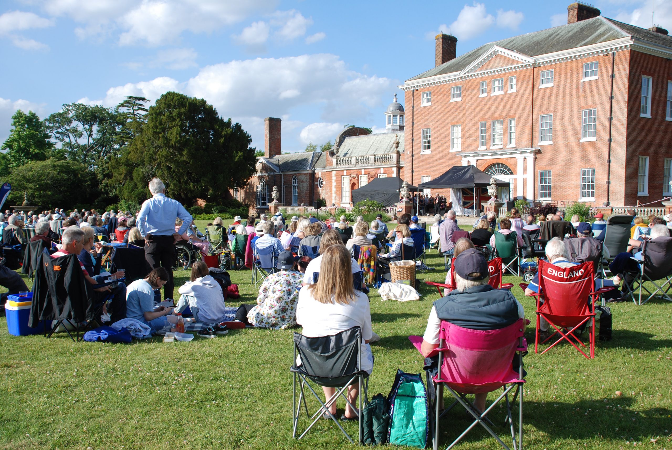 People sitting in the grounds of the National Trust’s Hatchlands Park House on a sunny day, enjoying Music in the Park. 