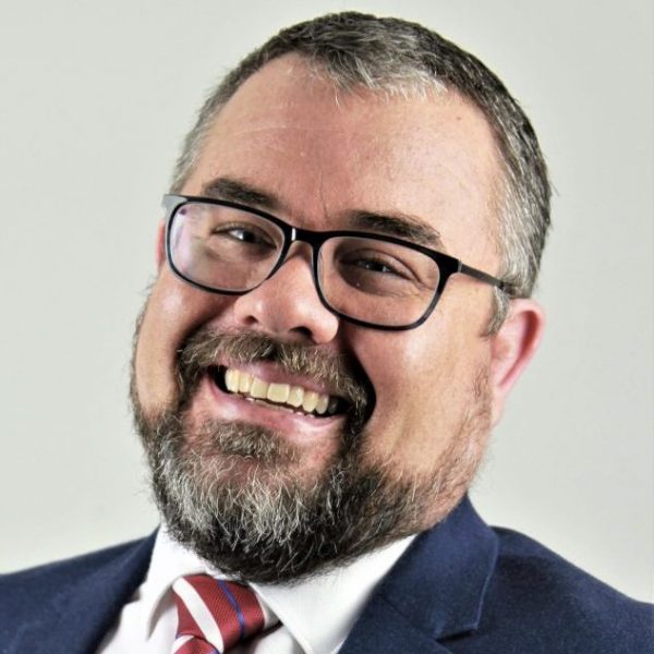 Craig with a big smile, wearing glasses, white shirt, tie and blue blazer.