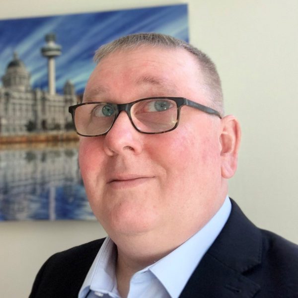Pete Hoey wearing glasses, blue shirt and black blazer with photo in the background of a building.