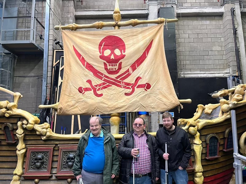3 men standing in front of the side of a pirate ship.