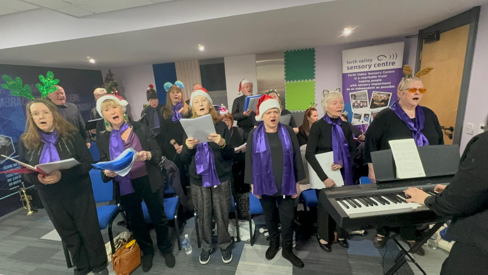 Sensory Singers choir performing, dressed in festive santa hats and headbands andaccompanied by a keyboard.