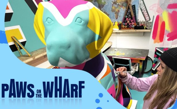 Graphic with the Paws on the Wharf logo and a front view image of an artist from duo Art+Believe painting a guide dog sculpture in a multi-coloured geometric design.