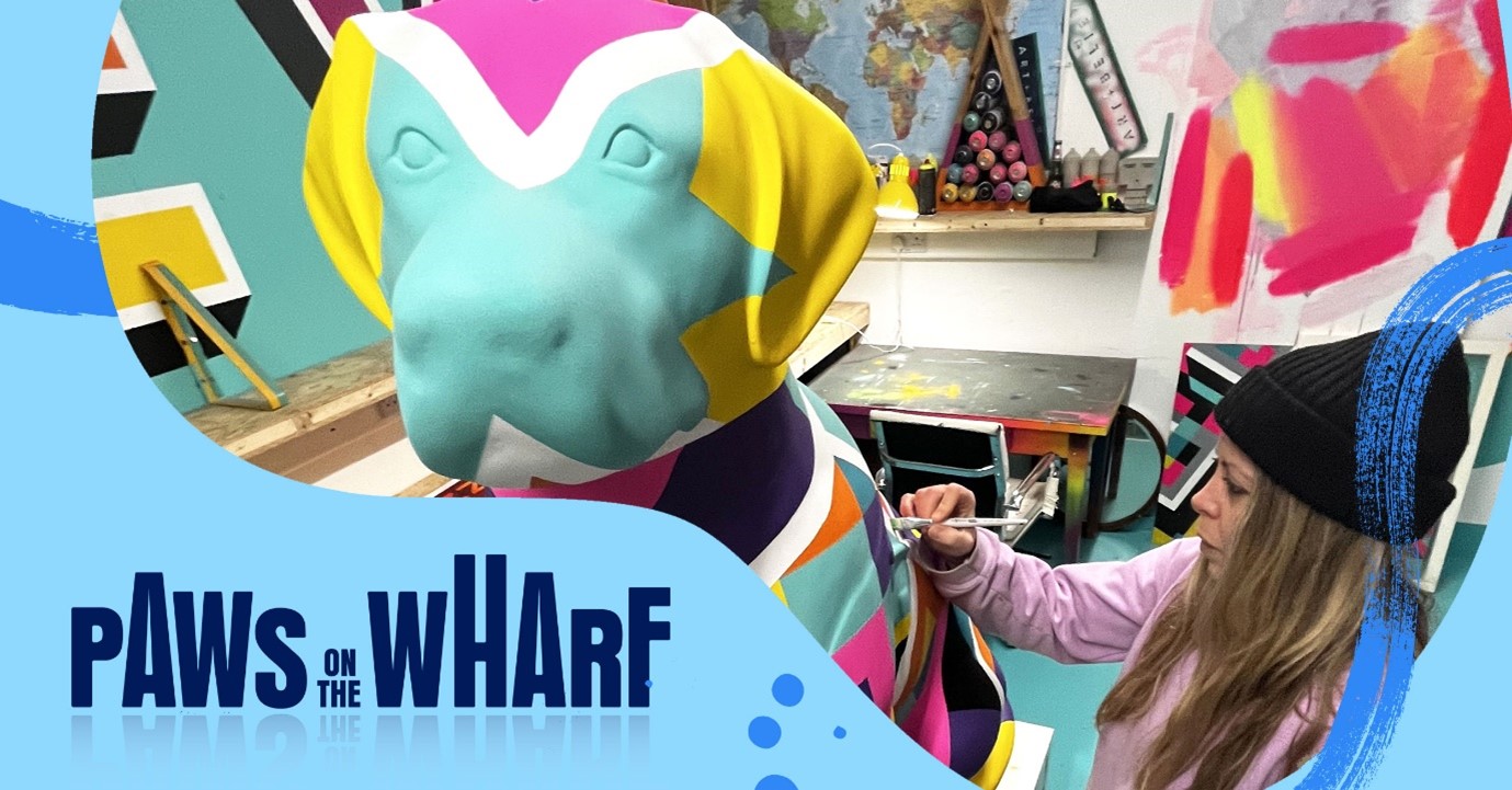 Graphic with the Paws on the Wharf logo and a front view image of an artist from duo Art+Believe painting a guide dog sculpture in a multi-coloured geometric design.