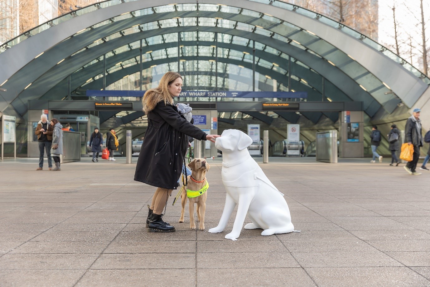 Guide dog owner with her yellow Labrador retriever guide dog standing next to a blank white guide dog sculpture in front of Canary Wharf tube station.