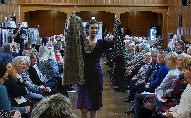 A model on the runway holds up 2 dresses. The audience is seated in rows on the right and left of the runway.