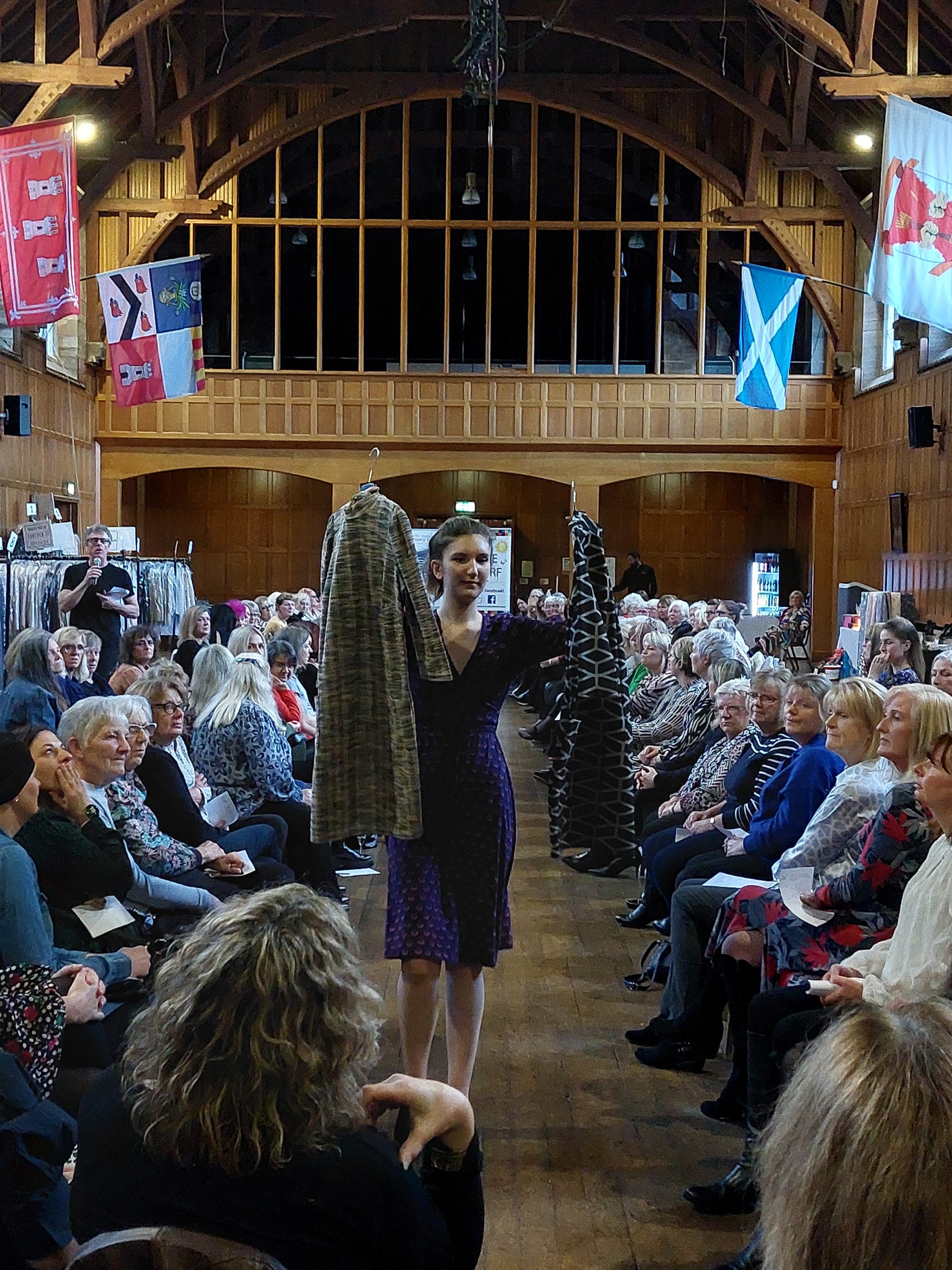A model on the runway holds up 2 dresses. The audience is seated in rows on the right and left of the runway.