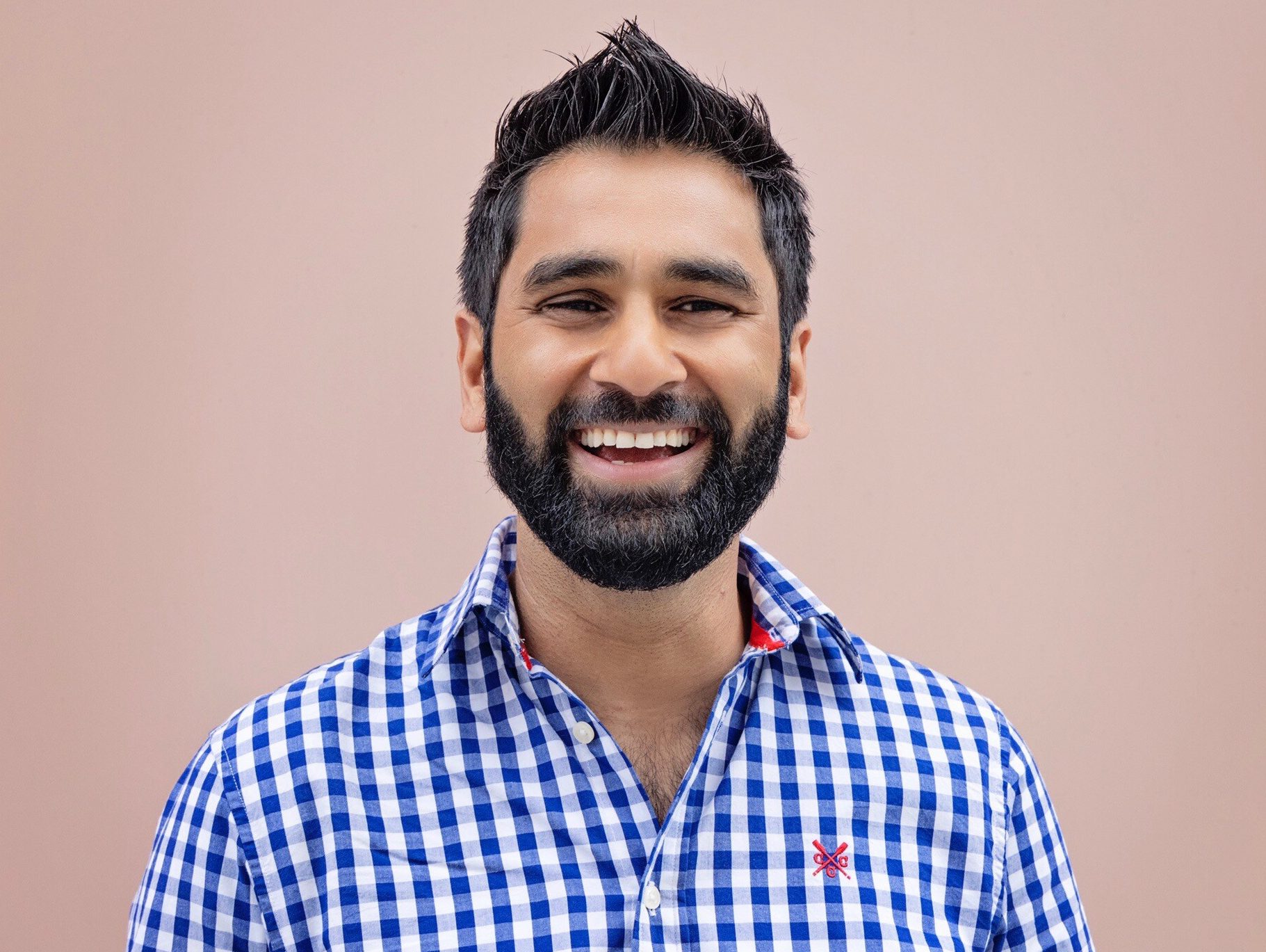 Dr Amit Patel has a beard and moustache. He is smiling, wearing a blue and white check shirt standing against a pink background.