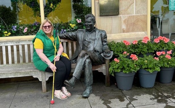 Amanda on a bench with her long yellow cane, she is sitting next a statue of a seated man