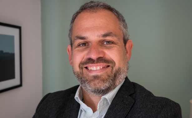 Ed Holloway smiling. He has a beard and moustache and is wearing a black blazer and shirt with top button open.