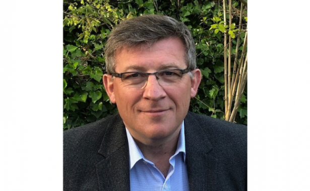 Matt Stringer wearing glasses, blue shirt and black blazer. There is leafy, green shrub in the background.