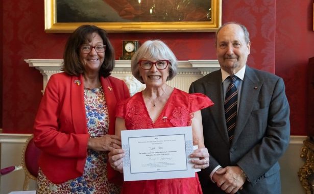 Photographer www.acumenimages.com. The picture shows three people smiling. Judith Potts is in the middle, holding a certificate and wearing a pink dress and an umbrella brooch. To her right is Michael Sternberg KC wearing a grey suit and to Judith’s right is Baroness Altmann, wearing a red jacket and a flowered dress.