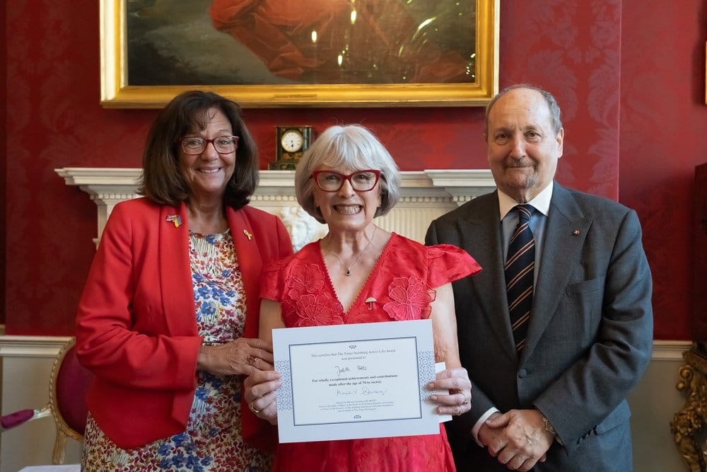 Photographer www.acumenimages.com. The picture shows three people smiling. Judith Potts is in the middle, holding a certificate and wearing a pink dress and an umbrella brooch. To her right is Michael Sternberg KC wearing a grey suit and to Judith’s right is Baroness Altmann, wearing a red jacket and a flowered dress.